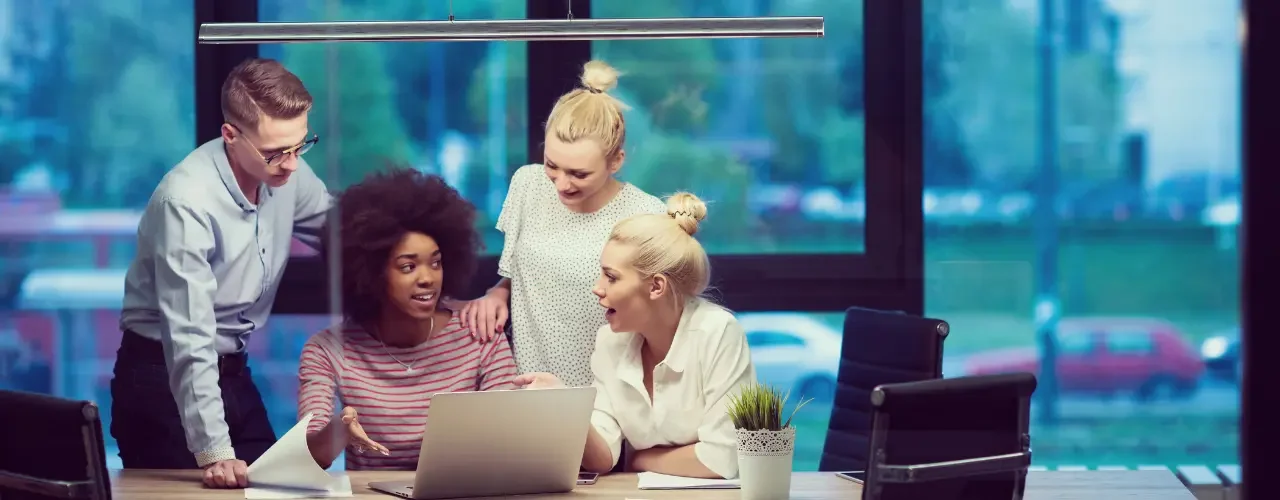 A group of colleagues sit at a desk and discuss something that is shown on a laptop.