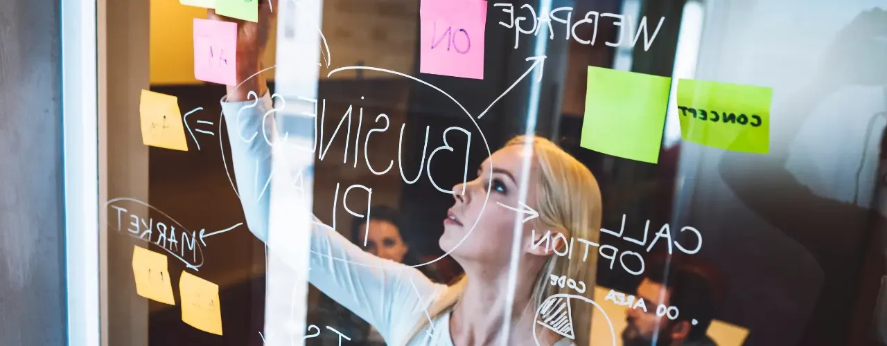 Success Story – Symbion 2 A woman is working on a whiteboard with sticky notes while her colleagues are watching.